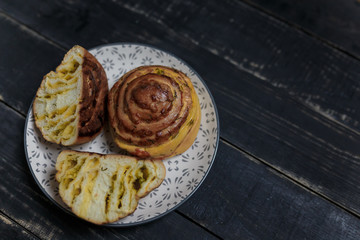 One whole and two halfs of cheese and dill bread bun on plate standing on black wooden background. Tasty unhealthy food. Diet, nutrition, eating habits concept. Place for text. 