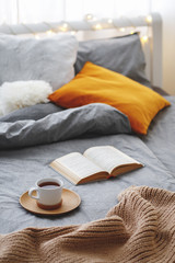 Cup of tea and book lie on the bed with gray linen, blanket, pillows, plaid and led garland light. Cozy winter home scene. Reading in bed.