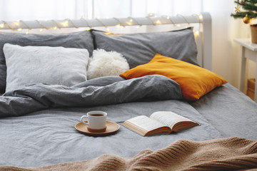 Cup of tea and book lie on the bed with gray linen, blanket, pillows, plaid and led garland light....