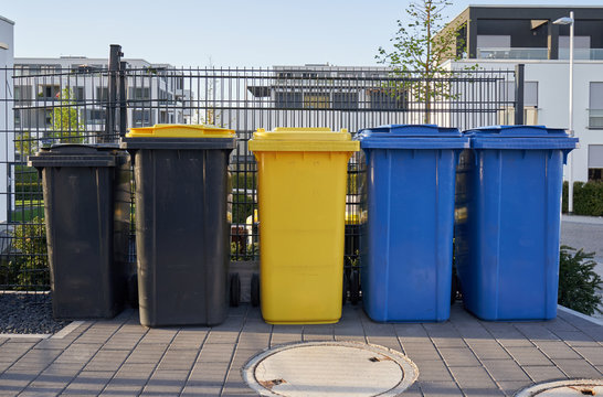 Different colors of garbage cans stand on the background of a fence made of mesh. Garbage containers for sorting garbage in a European city