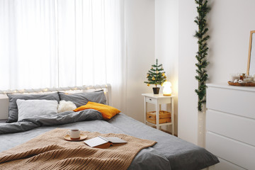 Christmas cozy winter home scene. New year interior decorations. Stylish cosy bedroom decor. Christmas tree on bedside table and bed with grey linen, blanket, pillows, knitted plaid, cup of tea, book.