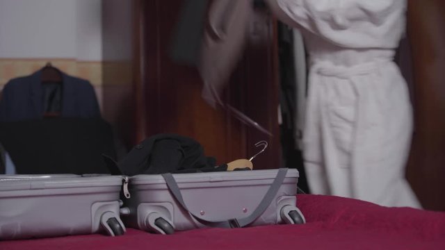 Young Caucasian unrecognizable woman in white bathrobe gathering clothes into suitcase. Nervous brunette girl throwing stuff in travel bag, closing it, and sitting on bed with back to the camera.