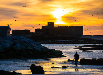 Silhouette of man walking away to the sea at sunset with ancient ruins on background in Essaouira,...