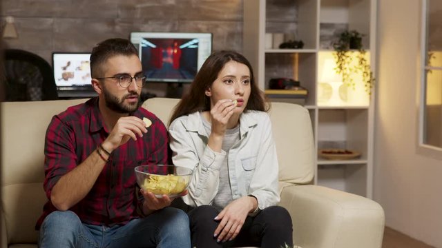 Caucasian young couple eating chips while watching TV
