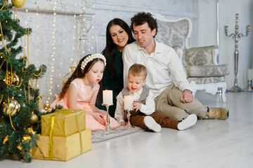 Young happy family at the Christmas tree. Children blow on candles and make wishes. Parents and their children celebrate the new year. New Year room with at home. Christmas good mood.