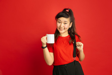 Naklejka premium Asian teenager's portrait isolated on red studio background. Beautiful female brunette model with long hair in casual style. Concept of human emotions, facial expression, sales, ad. Drinks tea, coffee
