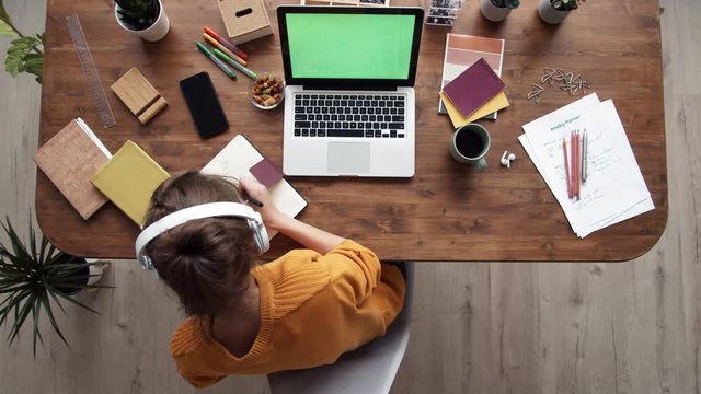 Time lapse of young female office worker sitting at desk, putting earphones on head, starting working on laptop and writing notes in notebook. Then she is going away