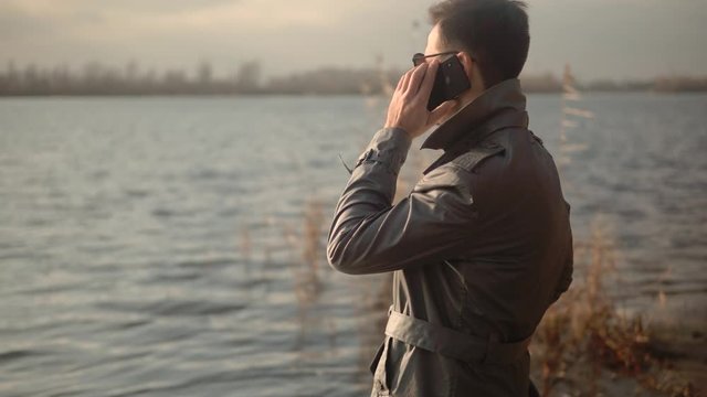 Handsome Man Talking By Smartphone Among Pond And Looking To Distance.Businessman At River Beach Using Mobile Phone And Enjoying Sunset.Beautiful Attractive Man Talking On Mobile Phone With Friend.