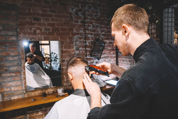 Young male barber shaves the client's head with a electric trimmer. Young bearded man getting haircut by hairdresser at barber shop. Advertising and barber shop concept