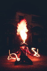 Man in red jester costume with double fire torch performing some action moment, spit fire up on top...
