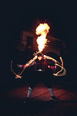 Man in red jester costume with double fire torch performing some action moment, spit fire up on top...