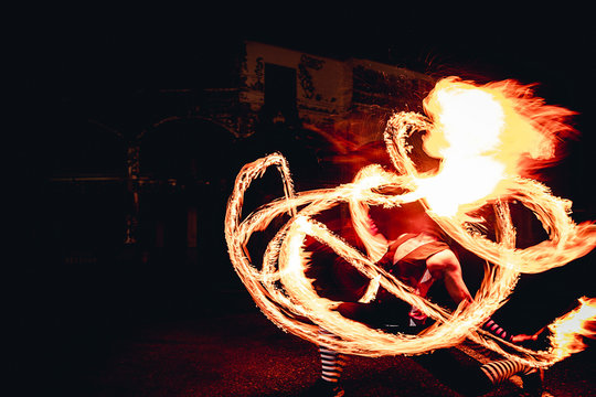 Man In Red Jester Costume With Double Fire Torch Performing Some Action Moment, Spit Fire Like Dragon Flame In The Dark, Long Exposure Style