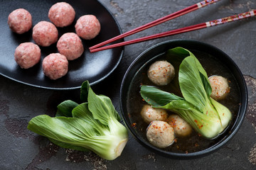 Miso soup with meatballs and pak-choi in a black bowl, studio shot on a brown stone surface with uncooked meatballs on a black plate
