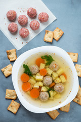 Soup with meatballs, potato, carrot and parsley. Flatlay over grey concrete surface with raw meatballs and crackers