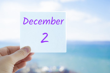 December 2nd. Hand holding sticker with text December 2 on the blurred background of the sea and sky. Copy space for text. Month in calendar concept