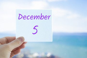 December 5th. Hand holding sticker with text December 5 on the blurred background of the sea and sky. Copy space for text. Month in calendar concept