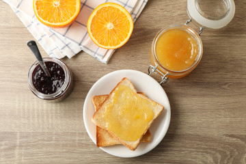 Toasts on plate, jam, towel and orange on wooden background, top view