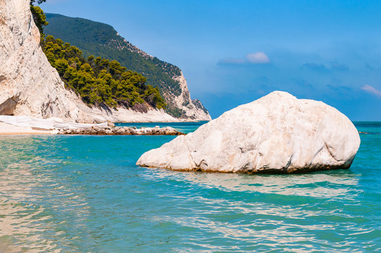Massive white boulder as a sea stack lying in water of Adriatic sea with beautiful coastline of Numana, Italy on the background. High massive white limestone rocky cliffs overgrown with trees