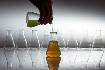 Hand pouring green in to orange liquid in scientific laboratory glass erlenmeyer flask with glassware equipment on reflective surface.