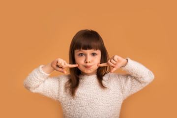 Harmful naughty child girl on an orange background grimaces and points fingers to her cheeks and...