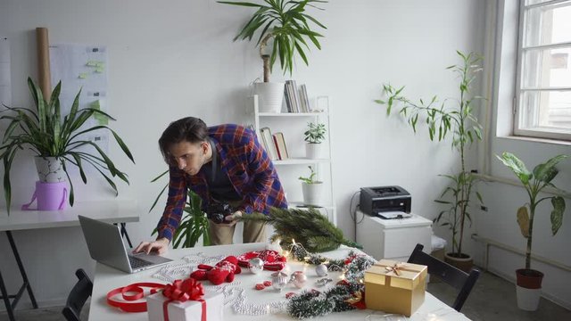 Medium shot of young male photographer arranging objects on table and taking Christmas flatlay images with camera, then checking pictures on laptop