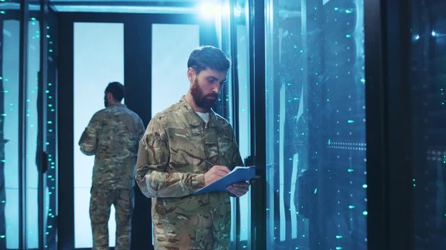 Caucasian military IT engineer monitoring a server cabinet with army officer, copying important data information writing down, cooperating on mission at data center.