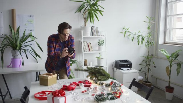Medium shot of young photographer or blogger arranging objects on table and taking Christmas flatlay images with camera from above