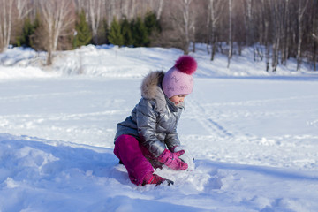 Fototapeta na wymiar Little girl in a silver jacket and a hat with a pompom walks in a winter park and rolls a big snowball