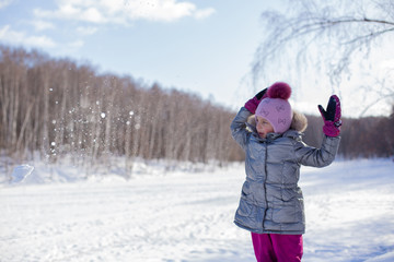 Fototapeta na wymiar Little girl in a silver jacket and a hat with a pompom walks in a winter park makes and throws large snowballs