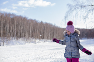 Fototapeta na wymiar Little girl in a silver jacket and a hat with a pompom walks in a winter park makes and throws large snowballs