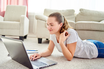 Portrait of happy smiling young woman lying on floor and using laptop at home, copy space