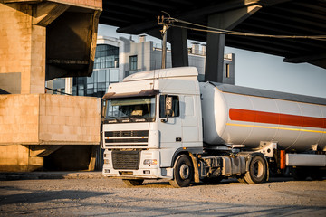 Truck with trailer, tank with flammable liquid, under a large bridge at the pier on the river bank, sunset light, white and red cars standing on gravel