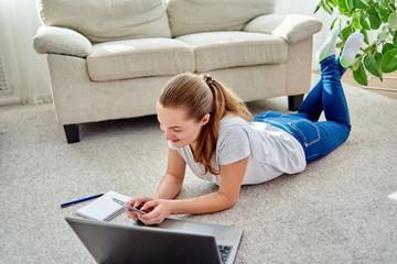 Portrait of happy smiling young woman lying on floor with laptop and using mobile phone at home, copy space