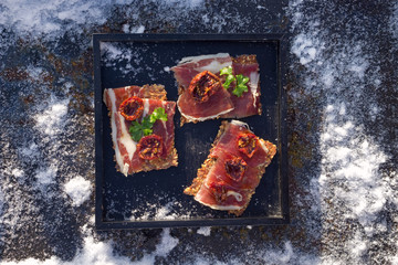  A black tray  with  Traditional  tapas    with  spanish cured meat, bread chips with seed,  sun dried tomato , parsley on background