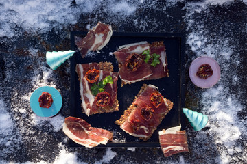  A black tray  with  Traditional  tapas    with  spanish cured meat, bread chips with seed,  sun dried tomato , parsley and  wooden plate and toy tree on background