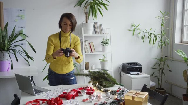 Medium shot of middle aged female photographer arranging objects on table and taking Christmas flatlay images with camera, then checking pictures on laptop