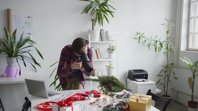 Professional male photographer arranging holiday objects on table and taking Christmas flatlay images with camera, then checking pictures on laptop computer
