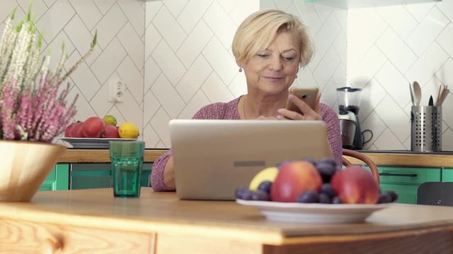 Senior woman using laptop and smartphone sitting in the kitchen