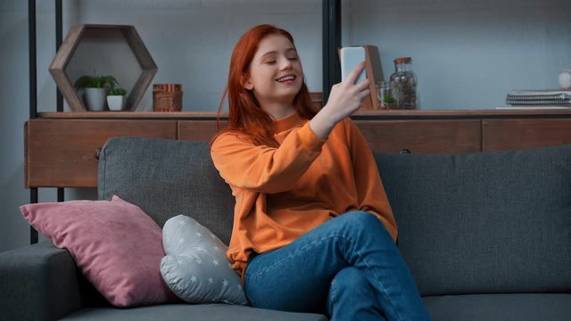 cheerful teenage girl sitting on sofa and taking selfie