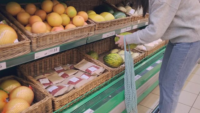 A young woman in a supermarket puts a melon in a mesh eco bag. The woman in the store uses a cotton mesh bag instead of plastic bags.