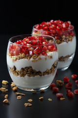  Yogurt with granola and pomegranate fruits in a glass cup on a black wooden table. Tasty dessert Healthy breakfast