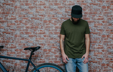 City portrait of handsome hipster guy with beard wearing a blank green military t-shirt and blank black cap standing on a brick wall background. Empty space for your logo or design. Mockup for print.