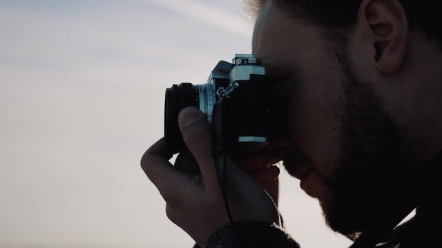 Close-up side view of young happy bearded creative photographer man taking a photo with vintage film camera slow motion.