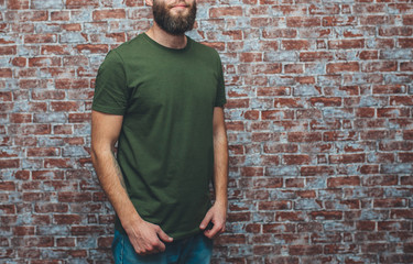 City portrait of handsome hipster guy with beard wearing a blank green military t-shirt standing on a brick wall background. Empty space for your logo or design. Mockup for print.