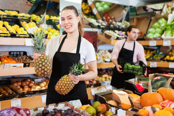 Positive saleswoman in apron offering fresh pineapples on the fruit store