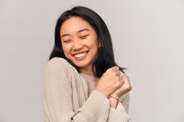 Satisfied girl presses hands to chest, happy to receive gift. Businesslike young woman Asian appearance with black hair and brown eyes dressed in knitted warm sweater stands isolated white background