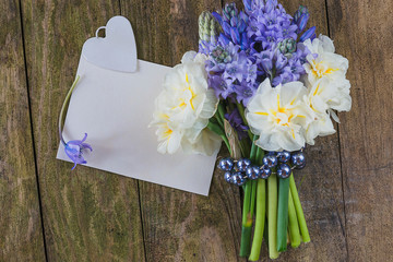 White paper, white heart and bouquet of white daffodils and blue hyacinths on the old wooden background. Copy space; flat lay