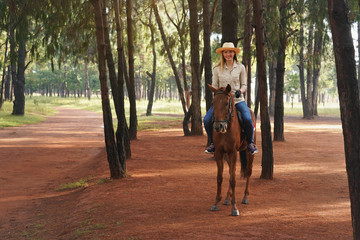 Fototapeta premium Young woman in shirt and straw hat, riding brown horse on park path, smiling, blurred trees in background