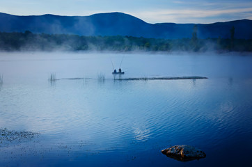 Early morning fog over the lake. Silhouettes of two men fishing in a boat. Landscape in trendy blue tones. Color of the year 2020, Classic Blue.