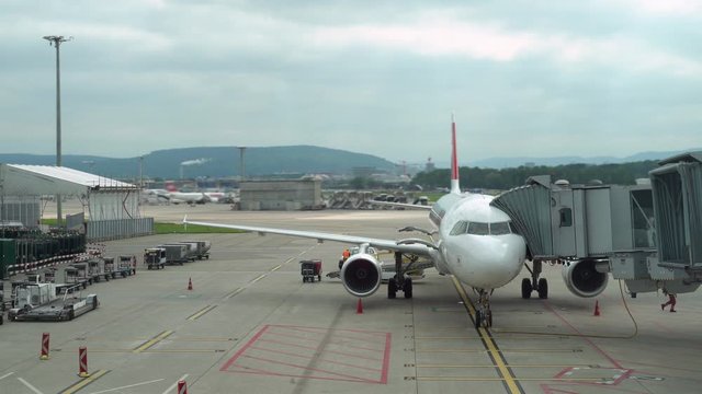Plane is parked at the airport near the terminal. Boarding tunnel attached to the plane. Airport buildings and other planes in the background. Close-up. Ultra HD stock footage